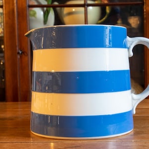 May include: A ceramic pitcher featuring a repeating horizontal striped pattern in bright blue and white. The vessel has a smooth, glossy finish, a wide cylindrical body, a curved handle on the right side, and a small pouring spout at the top rim.