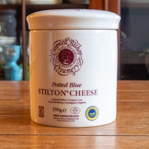 Może przedstawiać: Biały ceramiczny słoik z pokrywką, oznaczony jako "Potted Blue Stilton Cheese". Słoik posiada bordowe logo i tekst, o wadze 350g. Słoik jest cylindryczny i stoi na drewnianej powierzchni.