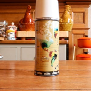 Könnte beinhalten: Eine Vintage-Thermosflasche mit weißem Deckel und zylindrischem Körper. Der Körper zeigt ein Kartendesign mit Kontinenten in Gelb-, Grün- und Blautönen. Auf der Basis der Flasche steht wiederholt das Wort "THERMOS".