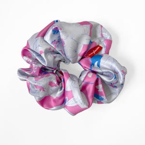 May include: A satin hair scrunchie featuring a vibrant pattern of pink, light blue, and white abstract shapes. A small red fabric tag with the white text marylouise is attached to the side of the gathered, circular hair accessory.