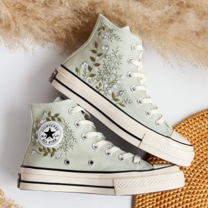 Op de afbeelding: Een paar lichtgroene Converse sneakers met hoge schacht en witte zolen. De sneakers zijn geborduurd met een bloemenpatroon en hebben een zwart "Converse All Star" logo aan de zijkant.