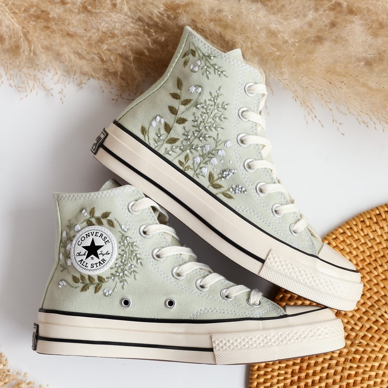 Op de afbeelding: Een paar lichtgroene Converse sneakers met hoge schacht en witte zolen. De sneakers zijn geborduurd met een bloemenpatroon en hebben een zwart "Converse All Star" logo aan de zijkant.