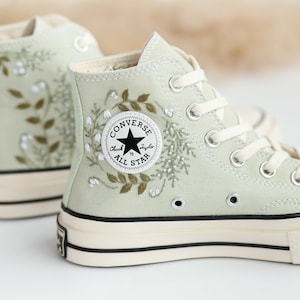 Op de afbeelding: Sagegroene hoge Converse sneakers met witte bloemenborduursel en een zwart Converse All Star logo.