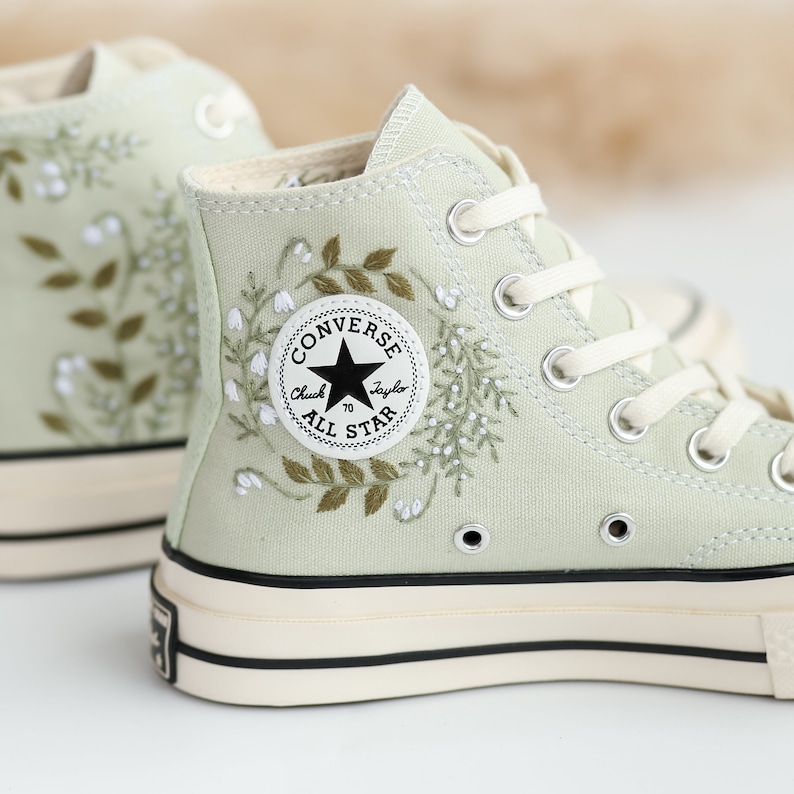 Op de afbeelding: Sagegroene hoge Converse sneakers met witte bloemenborduursel en een zwart Converse All Star logo.