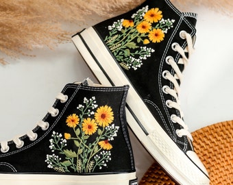 Può includere: Un paio di scarpe di tela nere alte con suole bianche. Le scarpe sono decorate con fiori gialli e foglie verdi ricamate.