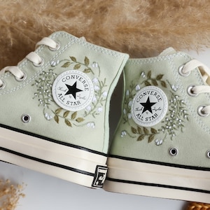Op de afbeelding: Een paar lichtgroene Converse sneakers met hoge schacht, geborduurd met witte bloemen en bladeren. Het Converse-logo is zichtbaar aan de zijkant van elke schoen.