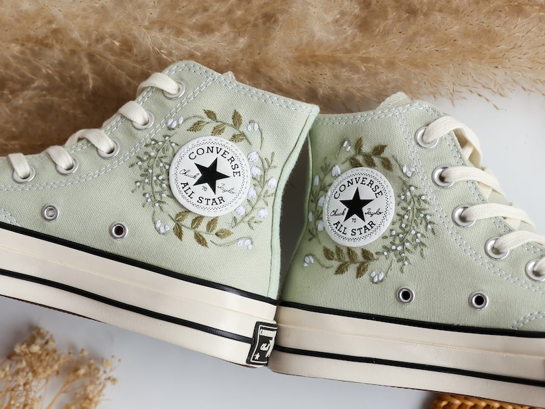 Op de afbeelding: Een paar lichtgroene Converse sneakers met hoge schacht, geborduurd met witte bloemen en bladeren. Het Converse-logo is zichtbaar aan de zijkant van elke schoen.