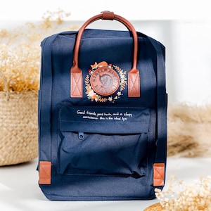May include: A navy blue backpack with brown leather straps and accents. The backpack has a large embroidered patch with a fox and the words "Fjällräven Kånken". The text "Good friends, good books, and a sleepy conscience: this is the ideal life" is embroidered below the patch.