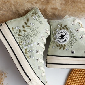 Op de afbeelding: Een paar lichtgroene Converse sneakers met hoge schacht en witte borduursels van bloemen en bladeren. De sneakers hebben een witte rubberen zool en zwarte stiksels. Het Converse-logo is zichtbaar aan de zijkant van de rechter schoen.