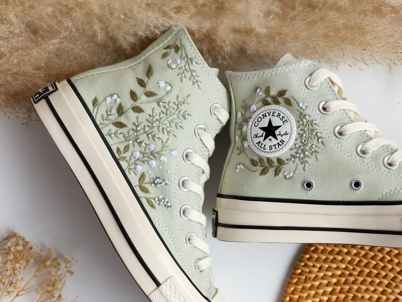 Op de afbeelding: Een paar lichtgroene Converse sneakers met hoge schacht en witte borduursels van bloemen en bladeren. De sneakers hebben een witte rubberen zool en zwarte stiksels. Het Converse-logo is zichtbaar aan de zijkant van de rechter schoen.