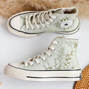 Op de afbeelding: Een paar lichtgroene high-top canvas sneakers met witte zolen en witte veters. De sneakers hebben een geborduurd bloemenpatroon met groene bladeren en witte bloemen. Het Converse All Star logo is zichtbaar aan de zijkant van de rechter schoen.