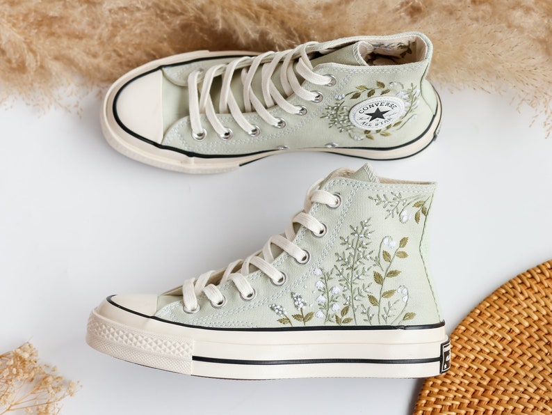 Op de afbeelding: Een paar lichtgroene high-top canvas sneakers met witte zolen en witte veters. De sneakers hebben een geborduurd bloemenpatroon met groene bladeren en witte bloemen. Het Converse All Star logo is zichtbaar aan de zijkant van de rechter schoen.