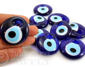May include: A group of ten blue glass evil eye charms with white and black centers. The charms are round and have a hole at the top for hanging.
