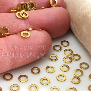 May include: A close-up of a pile of gold-colored jump rings. The jump rings are scattered on a white surface. The jump rings are small and round, with a diameter of about 1/4 inch.