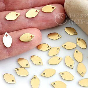 May include: A collection of 20 gold-coloured metal leaf-shaped charms with a hole at the top. The charms are scattered on a white surface.