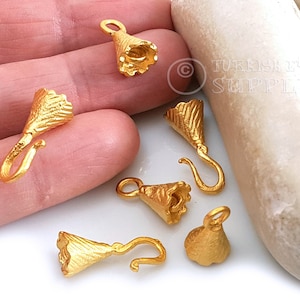 Puede incluir: Seis colgantes de metal dorado en forma de flor con un gancho en la parte inferior. Los colgantes se muestran en una mano y una piedra blanca.