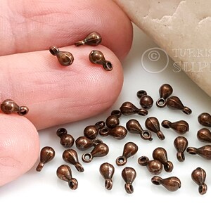 May include: A close-up of a pile of small, copper-colored teardrop-shaped beads with a loop at the top. The beads are scattered on a white surface.