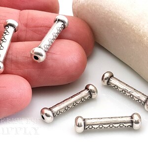 May include: Four silver metal beads with a decorative pattern. The beads are cylindrical and have rounded ends. The beads are arranged on a white surface.