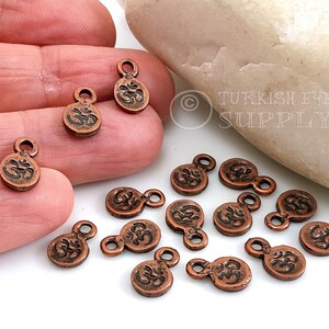 May include: A group of 13 copper-colored metal charms with an Om symbol engraved on each. The charms have a loop at the top for attaching to jewelry or other crafts.