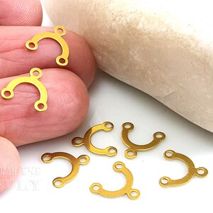 May include: Gold-toned metal connectors with three loops each, shaped like a rounded arch. The connectors are arranged on a hand and a light-colored surface.