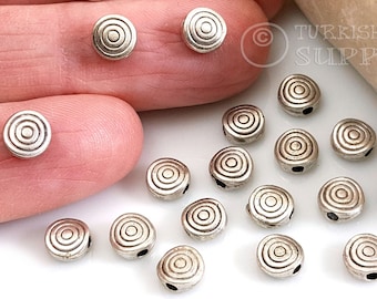 May include: Silver metal beads with a concentric circle design. The beads have a hole in the center for stringing. The beads are arranged on a white surface.