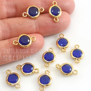 Pode incluir: Conectores de moldura dourados com cabochões de vidro azul escuro facetados. Os conectores têm pequenas argolas em cada lado para fixar a outros componentes de joalheria. A imagem inclui o texto "TURKISH EYE SUPPLY".