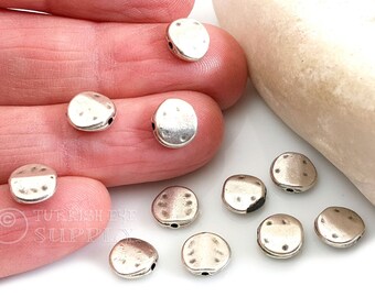 May include: A close-up of ten silver metal beads with a textured surface. The beads have two holes each and are scattered on a white surface.
