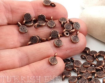 May include: A collection of small, copper-colored spiral charms. Each charm features a circular spiral design with a small loop for attaching to jewelry or other crafts. The charms are scattered across a surface, showcasing their intricate details.