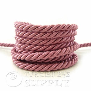 Puede incluir: Primer plano de una bobina de cordón trenzado de color rosa polvoriento. El cordón está hecho de un material suave y sedoso, ideal para proyectos de artesanía.
