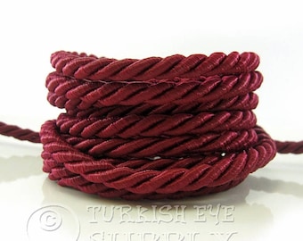 May include: A close-up of a spool of burgundy braided cord. The cord is twisted and piled on top of itself, creating a circular shape.
