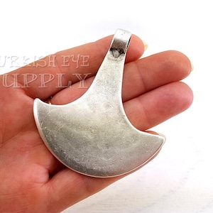 以下が含まれることがあります： 三日月型のデザインの、大きくて銀色のペンダント。ペンダントの上部にはチェーン用のループがあり、質感のある、古びた外観をしています。白い背景の中で、手で持たれています。