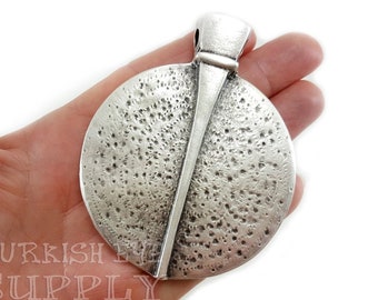 May include: A silver-toned pendant with a textured circular design and a vertical line down the center. The pendant has a loop at the top for attaching to a chain.