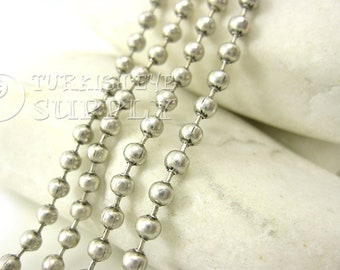 May include: Silver ball chain with a smooth, metallic finish. The chain is made of small, round beads that are linked together. The chain is lying on a white surface.