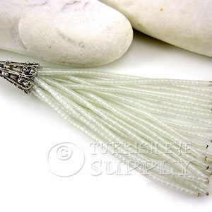 May include: A white beaded tassel with a silver metal top. The tassel is made of small, round beads that are strung together to create a long, flowing fringe.