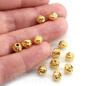 May include: A collection of small, round, gold-colored metal beads with a textured surface featuring recessed circular indentations. The beads have a central hole for threading and a polished, reflective finish, displayed in a group.