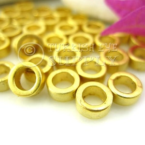 May include: A collection of small, gold-colored metal ring beads. The beads are cylindrical with a hole through the center. The beads are scattered on a white surface, with a few pink flower petals in the background. The beads have a matte finish.
