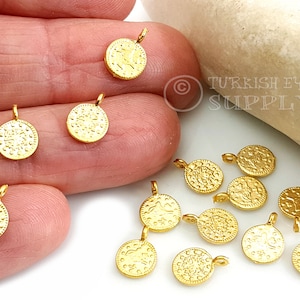 May include: A close-up of 12 gold-coloured coin charms with intricate designs. The charms have a loop at the top for attaching to jewellery or other crafts.