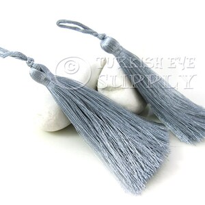 May include: Two silver gray tassel trim with a loop for attaching to a project. The tassels are on a white surface.