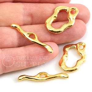 May include: Four gold-colored metal charms with irregular shapes and a hole for attaching to jewelry. The charms are on a hand, and the text "Turkish Eye Supply" is visible in the background.