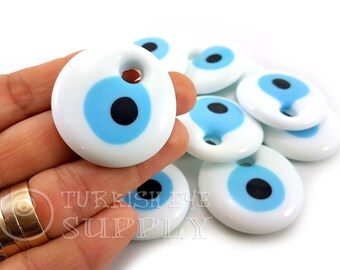 May include: A close-up of a hand holding a white ceramic evil eye charm with a blue and black eye design. The charm has a hole for hanging. There are several other charms in the background.