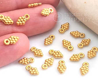 May include: Gold-toned metal grape cluster charms with a loop for attaching to jewelry or other crafts. The charms are arranged on a hand and a white surface.
