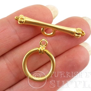 Può includere: Primo piano di due chiusure per gioielli color oro. Uno è una barra con estremità decorative e un anello, l'altro è un anello con un anello. Le chiusure sono probabilmente per collane o bracciali. Le chiusure sono tenute in una mano.