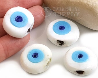 May include: Close-up of several white, round beads with a blue and light blue eye design. Each bead has a hole for stringing. The beads are likely for jewelry making or other crafts.