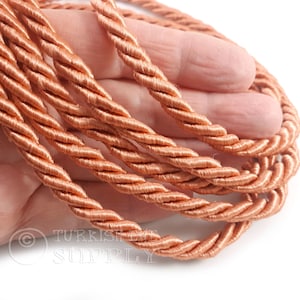 May include: A length of copper-colored twisted cord featuring a smooth, shiny finish and a multi-strand braided construction. The cord is coiled in several loops, displaying a consistent metallic sheen throughout its entire length.