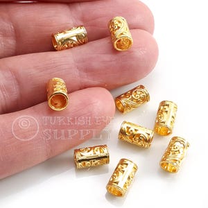 May include: Close-up of several gold-colored, cylindrical beads with intricate, openwork designs. The beads are scattered on a white surface, with some held in a hand. The beads have a shiny finish and are likely for jewelry making.