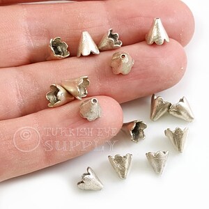 May include: Silver metal bead caps with a scalloped edge and a cone shape. The caps are scattered on a hand.