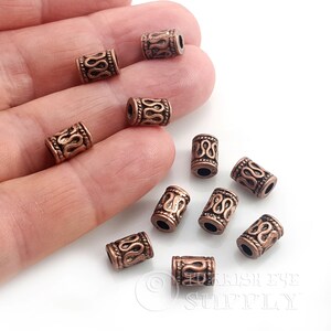 Peut inclure: Un ensemble de petites perles cylindriques couleur cuivre avec un motif ondulé en relief et des bords texturés. Chaque perle présente un trou central pour l'enfilage, avec une finition métallique et des détails sombres en creux.