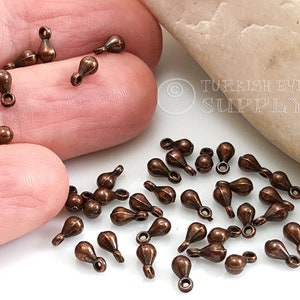 May include: A close-up of a pile of small, copper-colored teardrop-shaped charms with a loop at the top. The charms are scattered on a white surface.