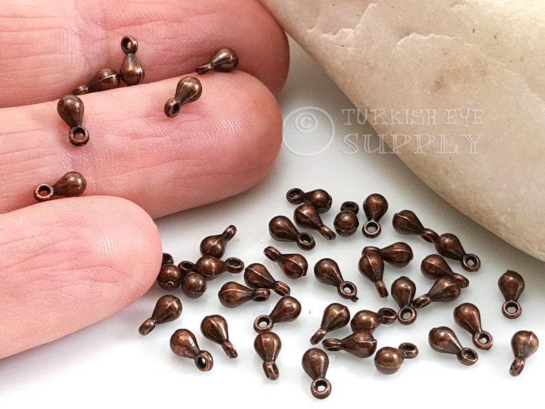 May include: A close-up of a pile of small, copper-colored teardrop-shaped charms with a loop at the top. The charms are scattered on a white surface.