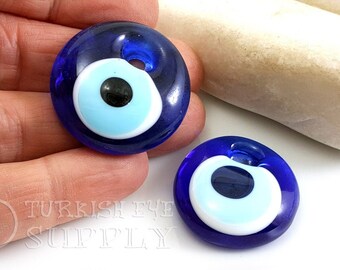 May include: Two round, dark blue glass pendants with a light blue and white eye design. Each pendant has a small hole at the top. The pendants are on a white surface with a light-colored stone in the background. The text "TURKISH EYE SUPPLY" is visible.